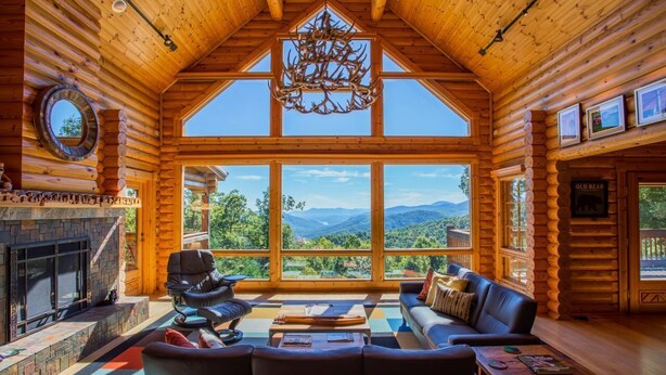 A log cabin interior with a fireplace, large windows, a chandelier, and a view of the mountains. - At Last Lodge | Pet Friendly Log Cabin with Hot Tub, Grill + VIEWS! (Old Fort)