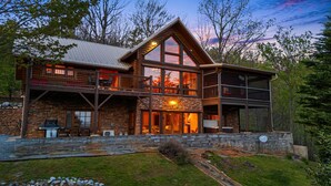 A two-story log cabin with a stone foundation, large windows, and a balcony. - At Last Lodge | Pet Friendly Log Cabin with Hot Tub, Grill + VIEWS! (Old Fort)