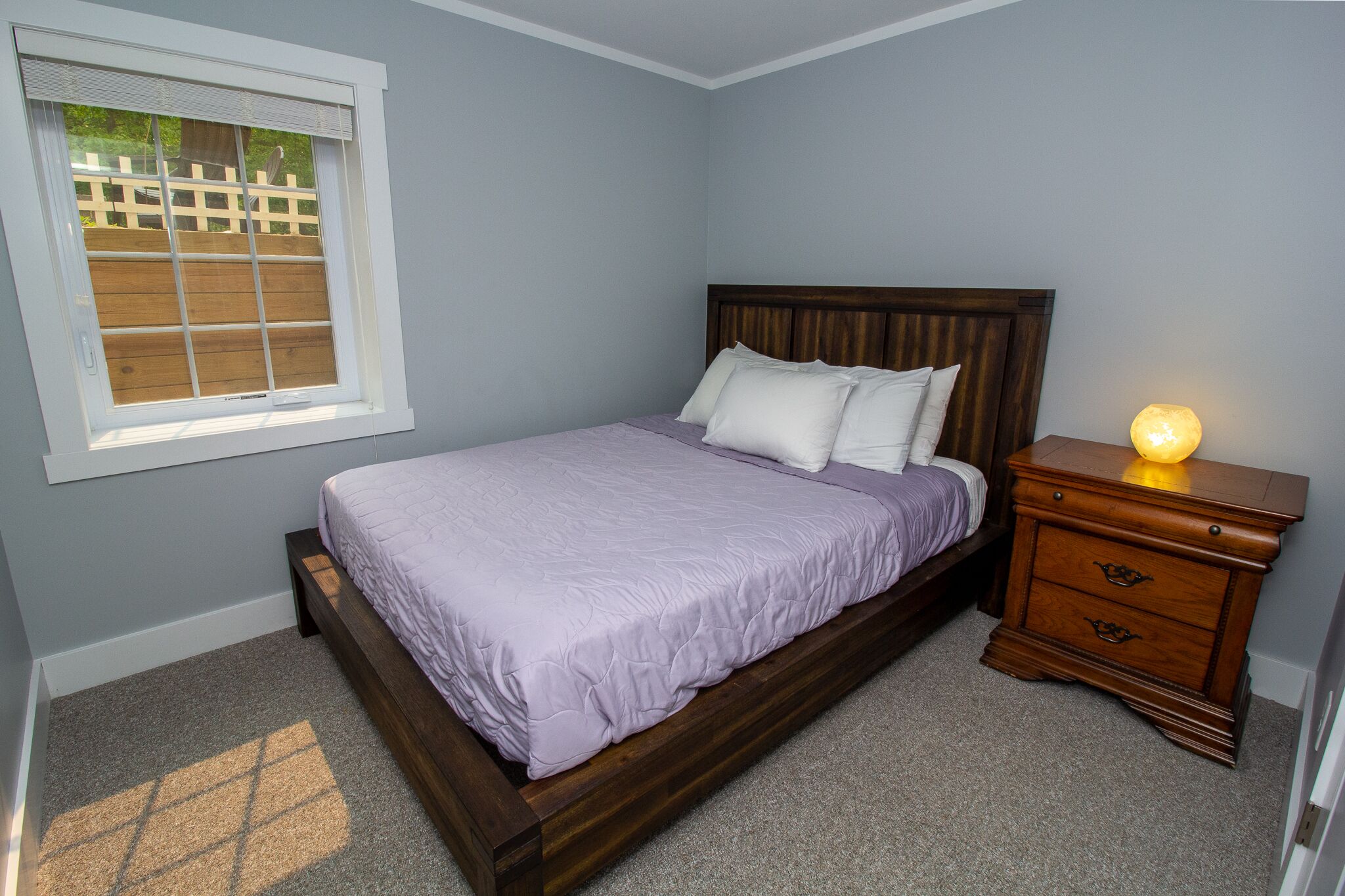 Un dormitorio con una cama de madera, una mesita de noche con una lámpara y una ventana que da a una valla de madera.