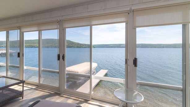 Ein Zimmer mit großen Fenstern mit Seeblick, eine Terrasse mit Liegestuhl und einem kleinen Tisch.