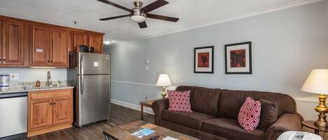 A compact living space with a kitchenette, a ceiling fan, a brown sofa with patterned cushions, a coffee table, and a lamp.