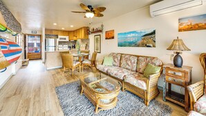 A spacious living area with a sofa, coffee table, and wicker chairs. There is a ceiling fan, kitchen area, and artwork on the walls. - Pristine Lihue Condo w/ Balcony: Walk to Beach! (Lihue)