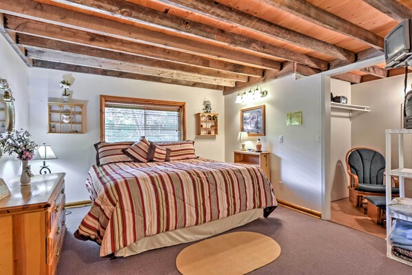 Un dormitorio con techo de madera, una cama con ropa de cama a rayas, una mesita de noche, una lámpara, una silla y un televisor.