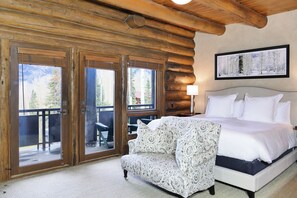 A log cabin room with a bed, patterned armchair, and large windows with a view of mountains.