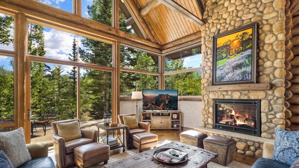 A spacious living room with a stone fireplace, large windows, and a television. - ALPEN RIDGE- Elevated Luxury Estate with Slopeside Views and Access (Mountain Village)