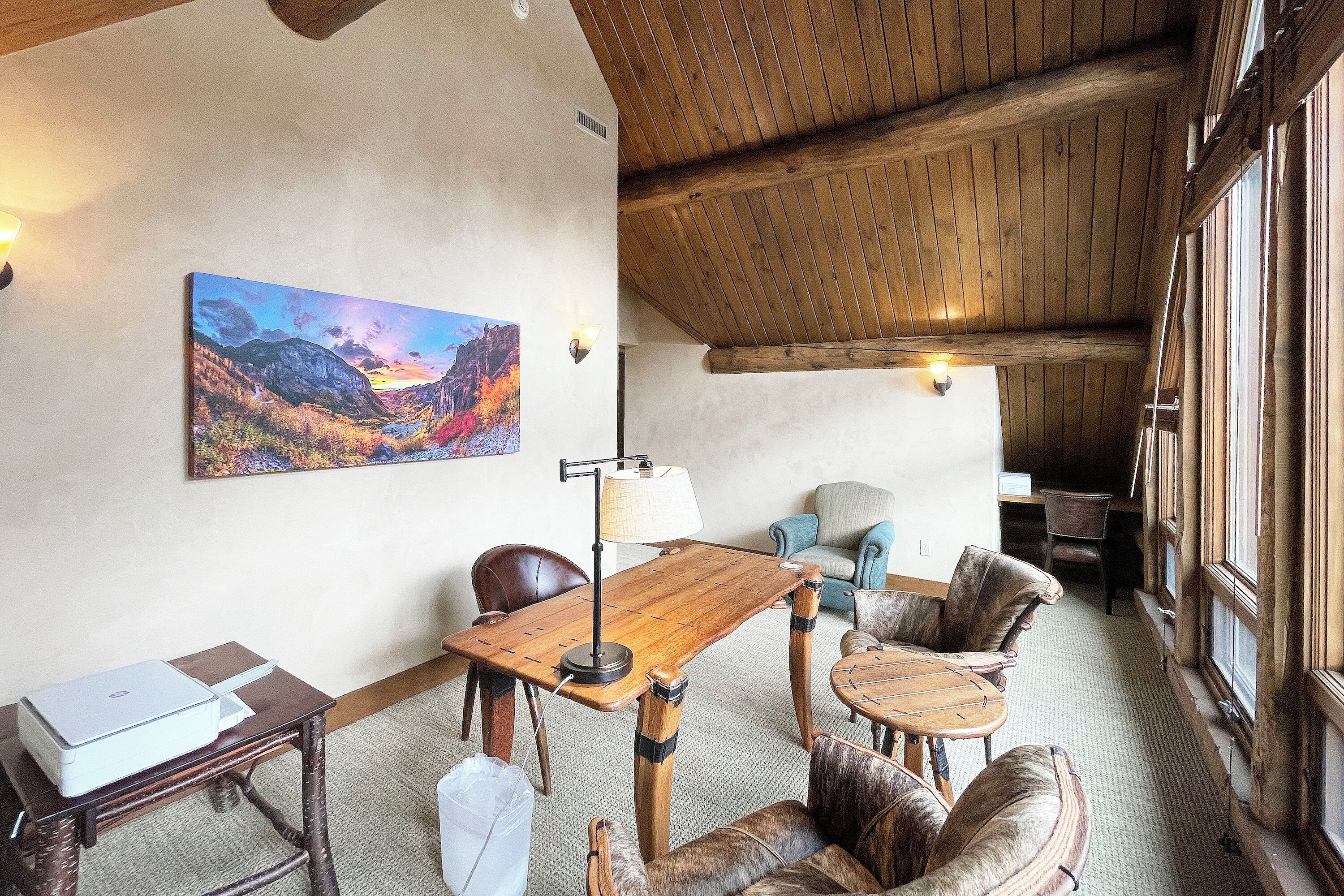 A cozy room with a wooden ceiling, a painting of a landscape, a table with a lamp, and leather chairs.