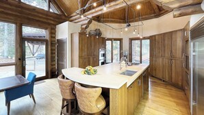 A modern kitchen with a large island, wooden cabinetry, and a dining area with blue chairs. - ALPEN RIDGE- Elevated Luxury Estate with Slopeside Views and Access (Mountain Village)
