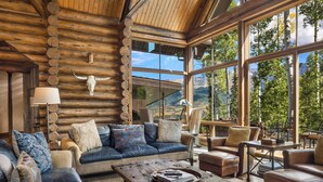 A log cabin interior with a large window offering a mountain view, leather furniture, and a rustic wooden coffee table. - ALPEN RIDGE- Elevated Luxury Estate with Slopeside Views and Access (Mountain Village)