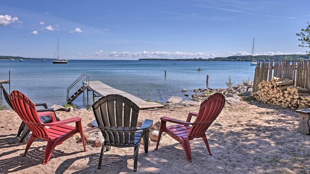 A beach with two Adirondack chairs, a wooden dock, and a stack of firewood. - 1/2 Mi to Dtwn! Suttons Bay Cottage w/ Fire Pit (Suttons Bay)
