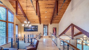 A spacious loft with high ceilings, wooden beams, a glass railing, and a view of the outdoors.