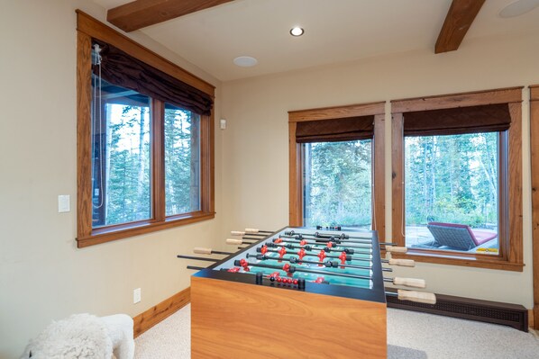 A foosball table in a room with large windows, wooden beams, and a view of trees.