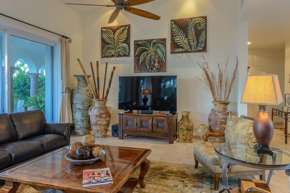 A living room with a leather sofa, a glass coffee table, a TV, decorative vases, and artwork on the wall. - Luxury Villa Steps to Calm Palmilla Swimming Beach w/ Pool, Hot Tub & Ocean View (Cabo San Lucas)