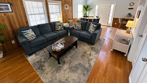 A living room with a dark gray sectional sofa, a wooden floor, a ceiling fan, and a dining area with a table and chairs. - KD108- Near the Beach- 4 BRs, Just a Short Stroll to the Beach, Dog Friendly! (Kill Devil Hills)
