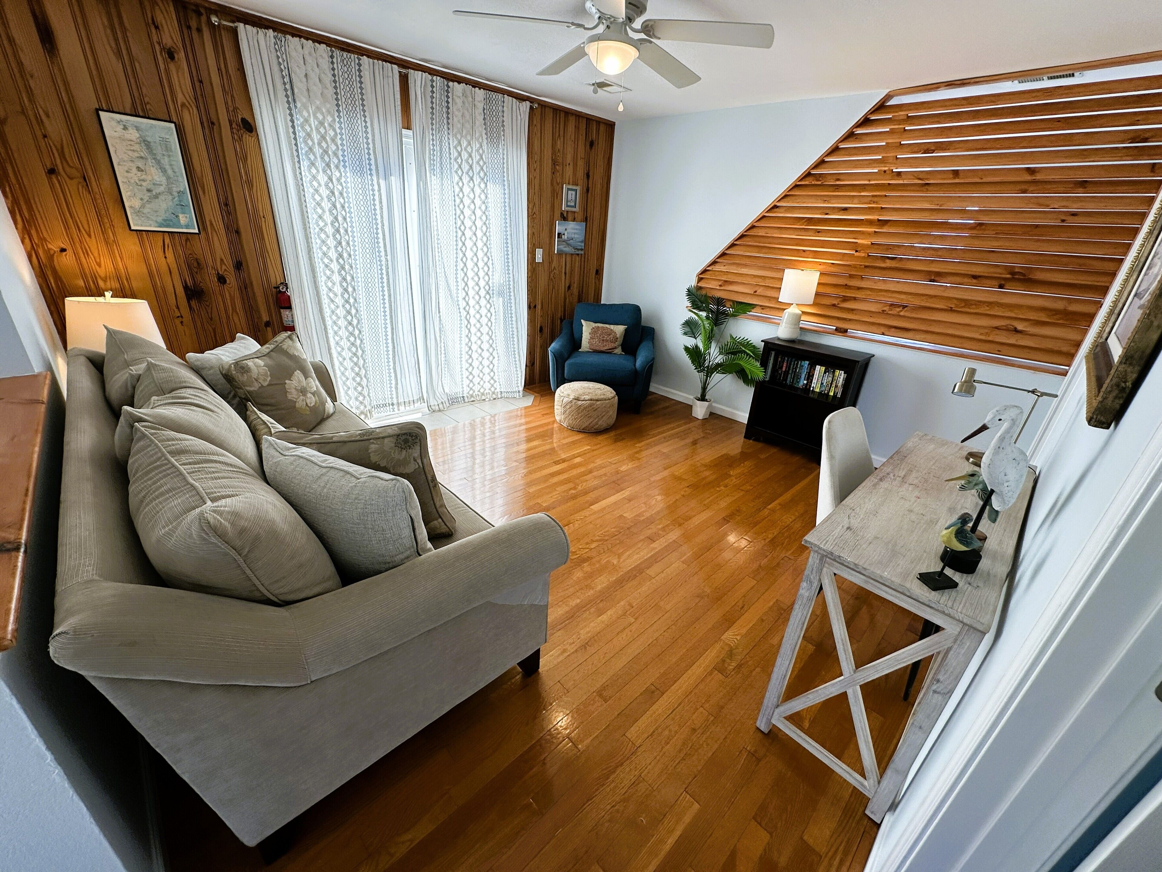 A living room with a sofa, a chair, a small table, a bookshelf, a plant, and a ceiling fan.