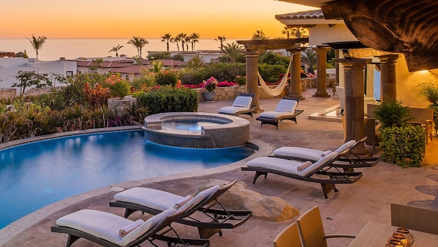 A pool area with lounge chairs and a hot tub, surrounded by plants and palm trees, overlooking a body of water.
