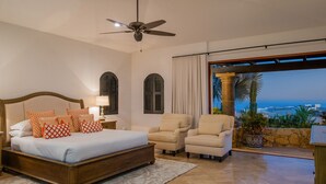 A bedroom with a large bed, two armchairs, a ceiling fan, and a view of the ocean.