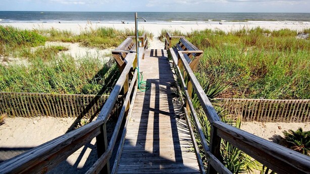 Ein hölzerner Steg führt zu einem Strand mit sandigem Ufer und grasbewachsenen Dünen.