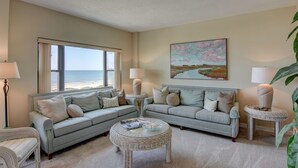 Una sala de estar con vista a la playa, un cuadro grande y dos mesitas de noche con lámparas.