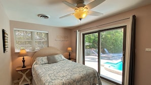 A bedroom with a bed, two bedside lamps, a ceiling fan, a window with blinds, and a view of an outdoor swimming pool.