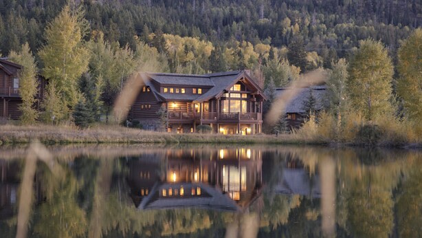 A log cabin with a reflection in a calm lake, surrounded by forested hills. - Salmonfly Lodge Remodeled Teton Springs Log Cabin in Victor w/ Hot Tub & A/C (Victor)