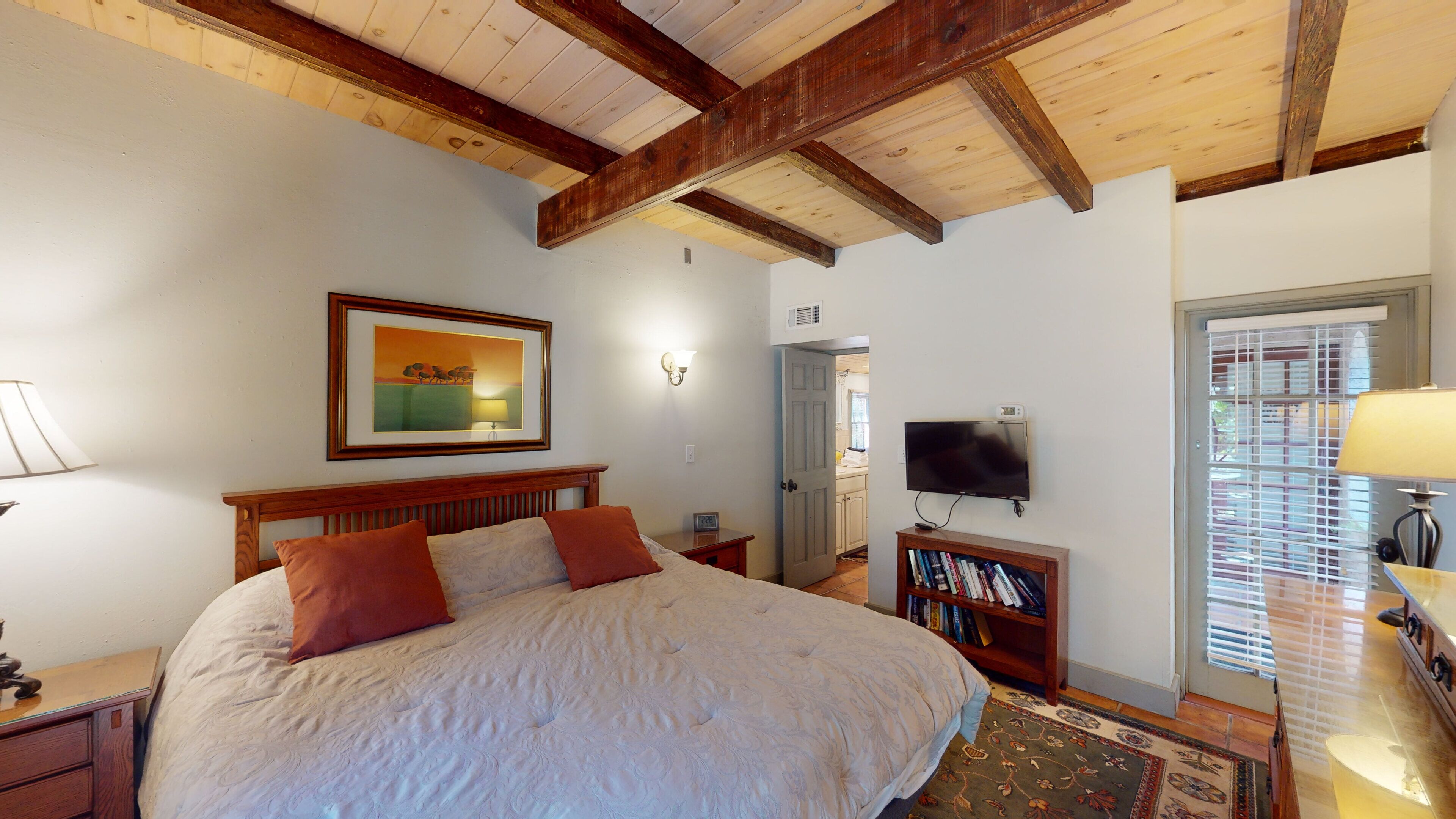 Ein Schlafzimmer mit einem großen Bett, Holzbalken, einem gerahmten Bild, einem Fernseher, einem Bücherregal und einer Tür zum Badezimmer.