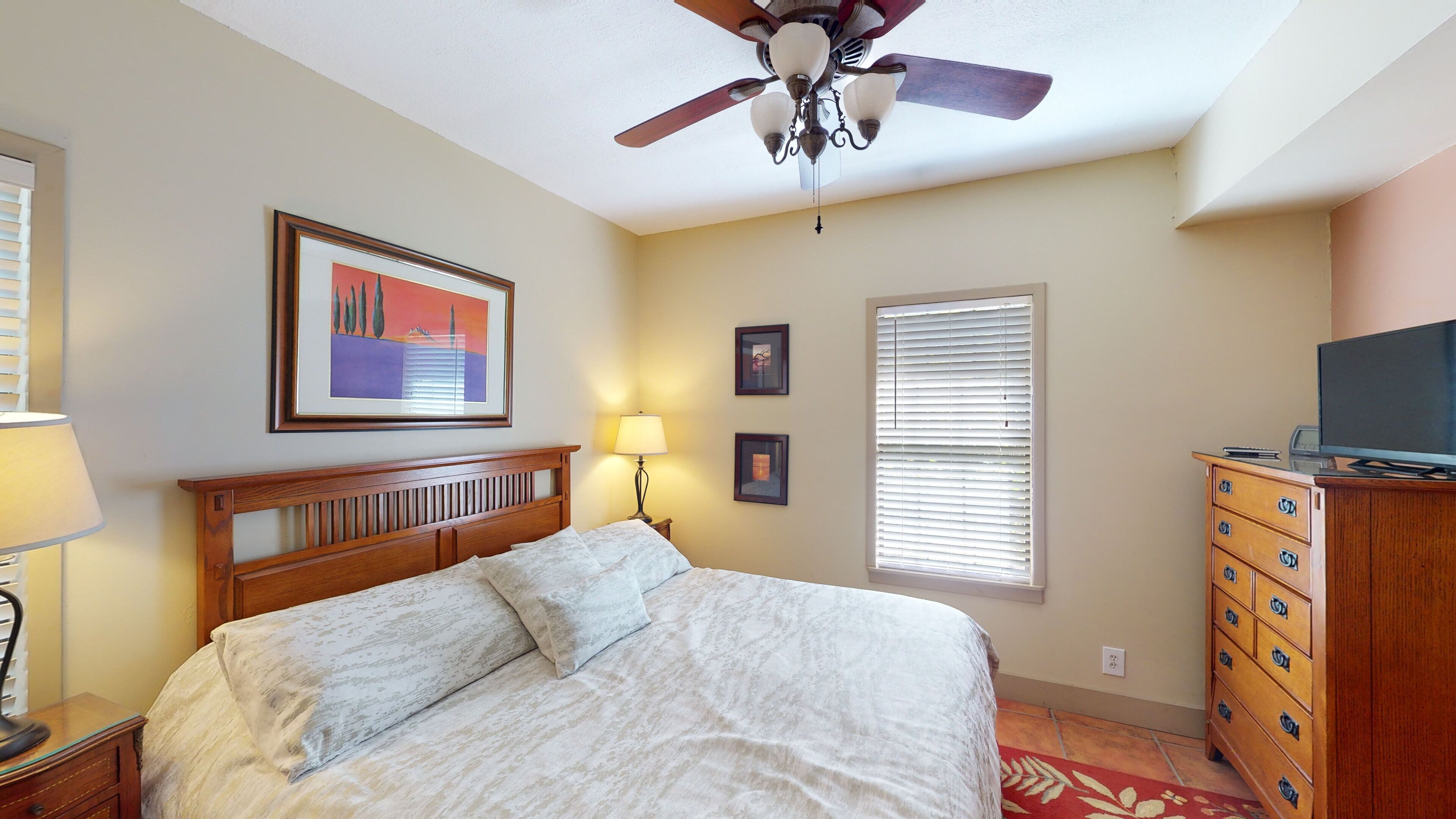 A bedroom with a bed, a ceiling fan, a nightstand with a lamp, a dresser, and a television.