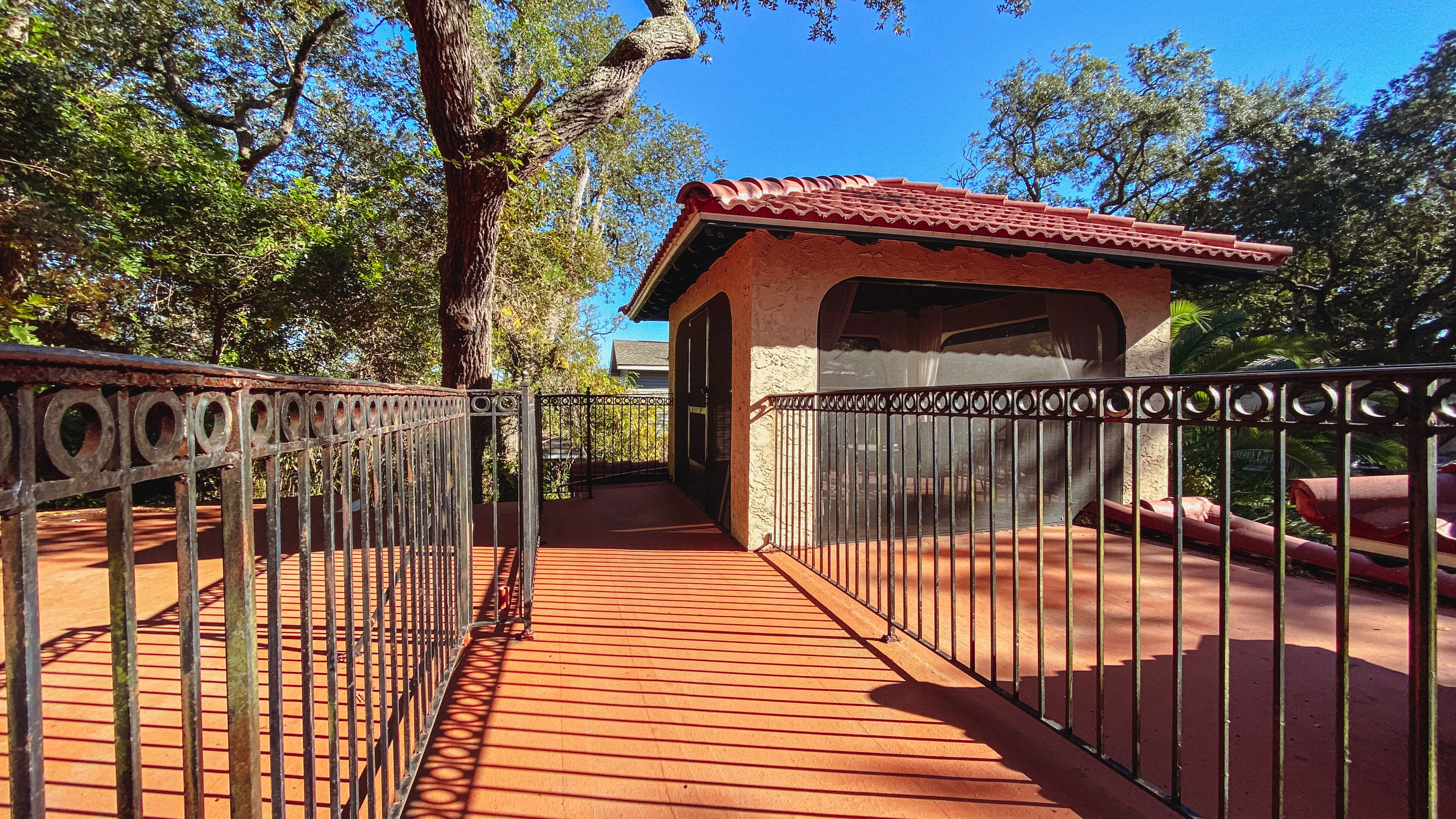A wooden pathway leading to a building with a red-tiled roof, surrounded by trees and a metal fence.