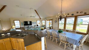 Una cocina amplia con una isla grande, armarios de madera y un televisor integrado. Hay un comedor con una mesa y sillas a cuadros, y un candelabro encima.
