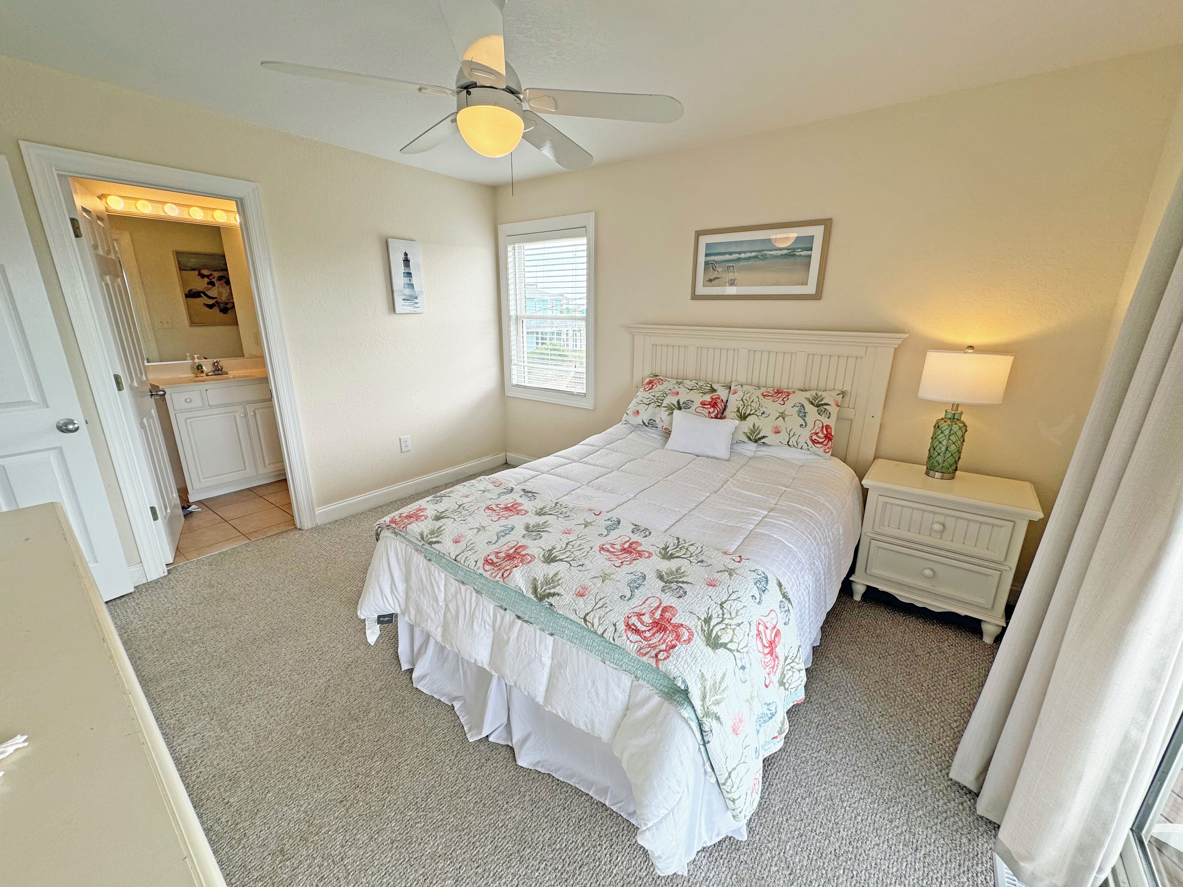 A bedroom with a bed, a nightstand, a ceiling fan, a window with blinds, a door leading to a bathroom, and a wall-mounted picture.