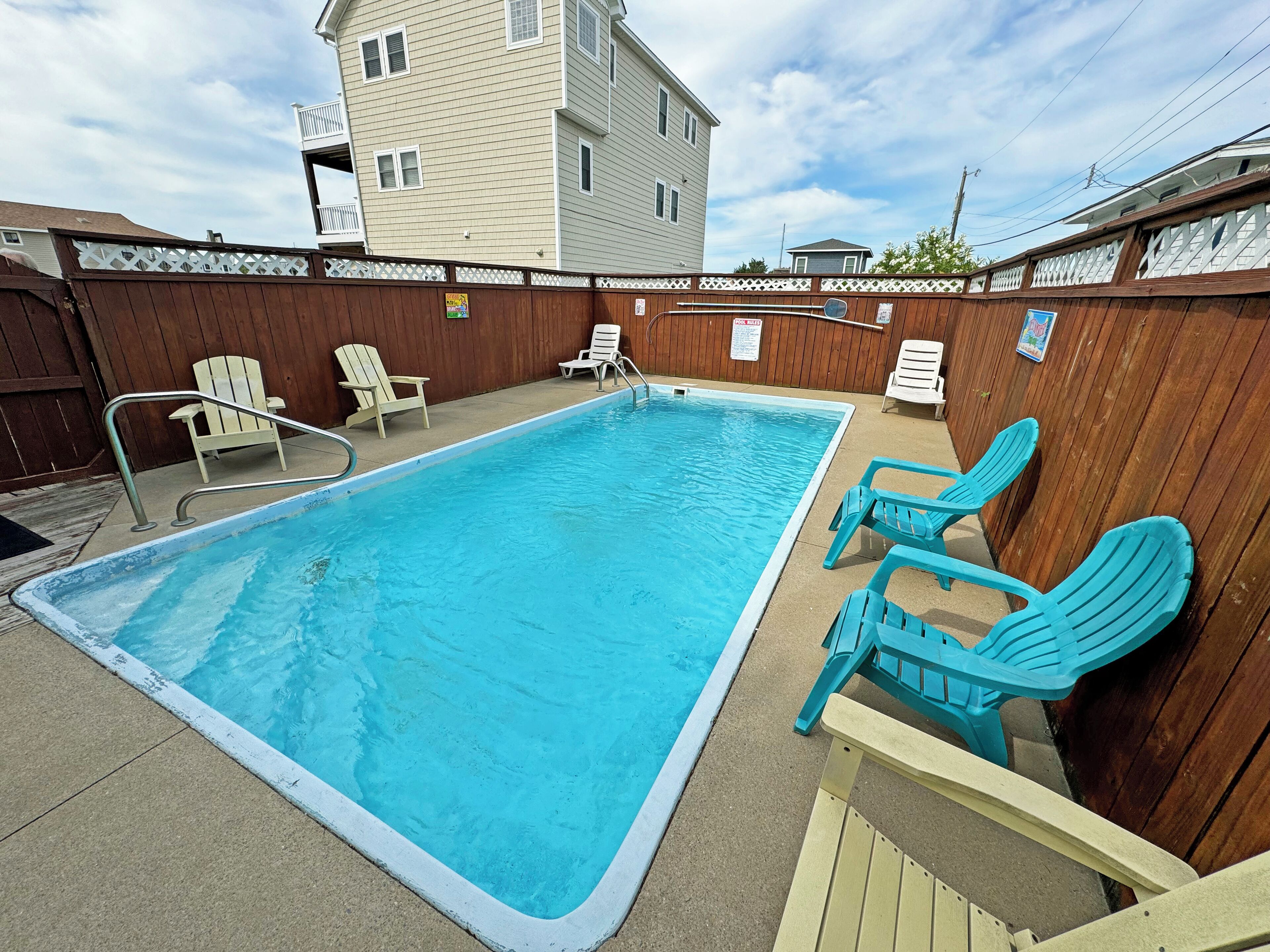 A rectangular swimming pool with a wooden deck, surrounded by a wooden fence and white chairs.