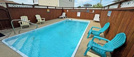 A rectangular swimming pool with a wooden deck, surrounded by a wooden fence and white chairs.