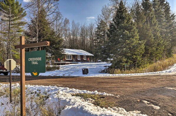 A wooden sign for Creekside Trail, a cabin with a snow-covered roof, and a forested area.