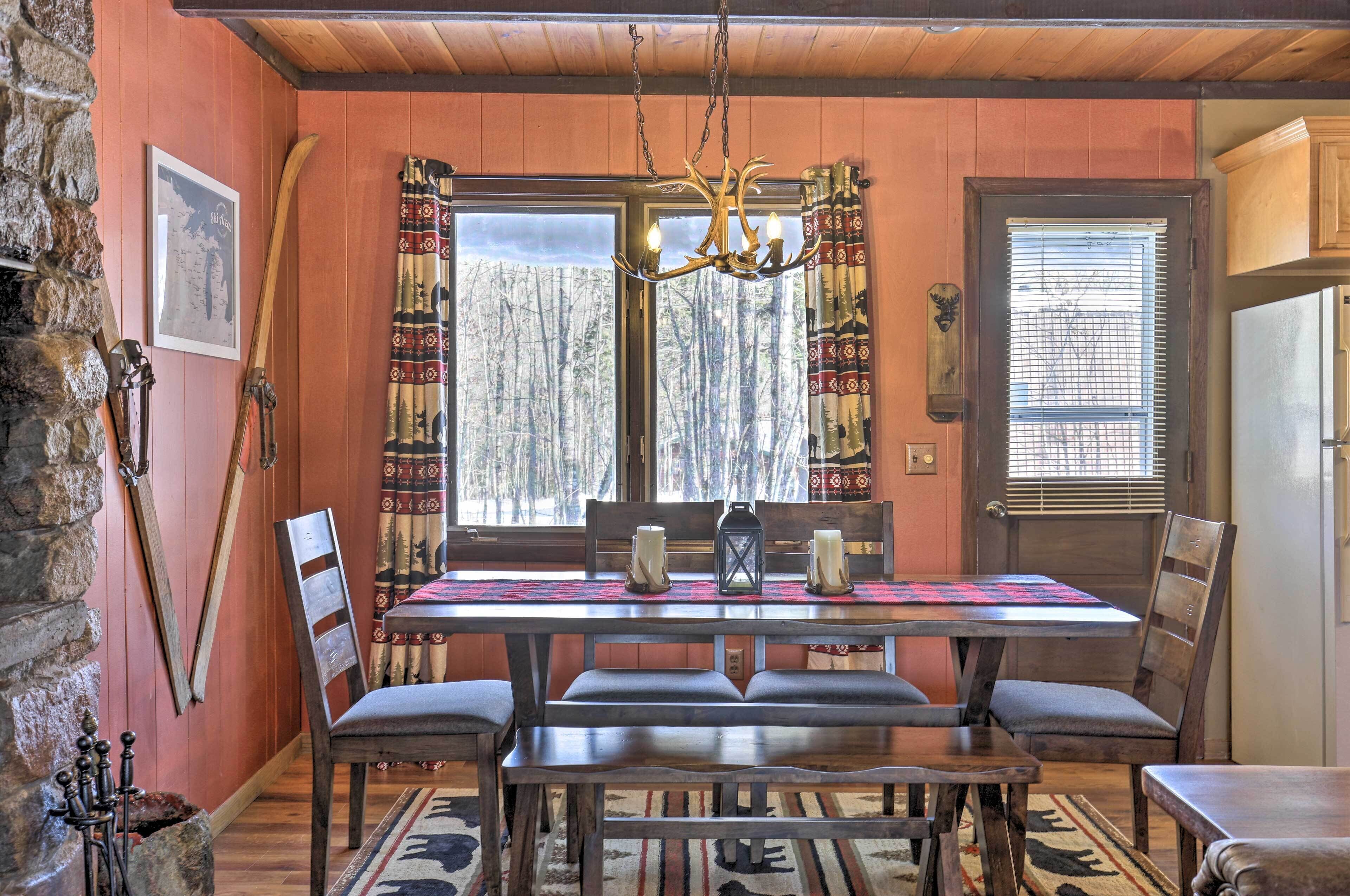 Un coin repas avec une table en bois, des chaises, un lustre et une vue sur les arbres à travers de grandes fenêtres.