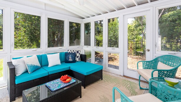 Eine überdachte Terrasse mit einem schwarzen Rattan-Sofa, türkisfarbenen Kissen und einem Glastisch.