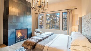 A bedroom with a fireplace, large windows, a chandelier, and a bed with a headboard. - PETRA DOMUS- SKI PASSES! Luxury Stone House w/ Hot Tub, Guest House (Mountain Village)