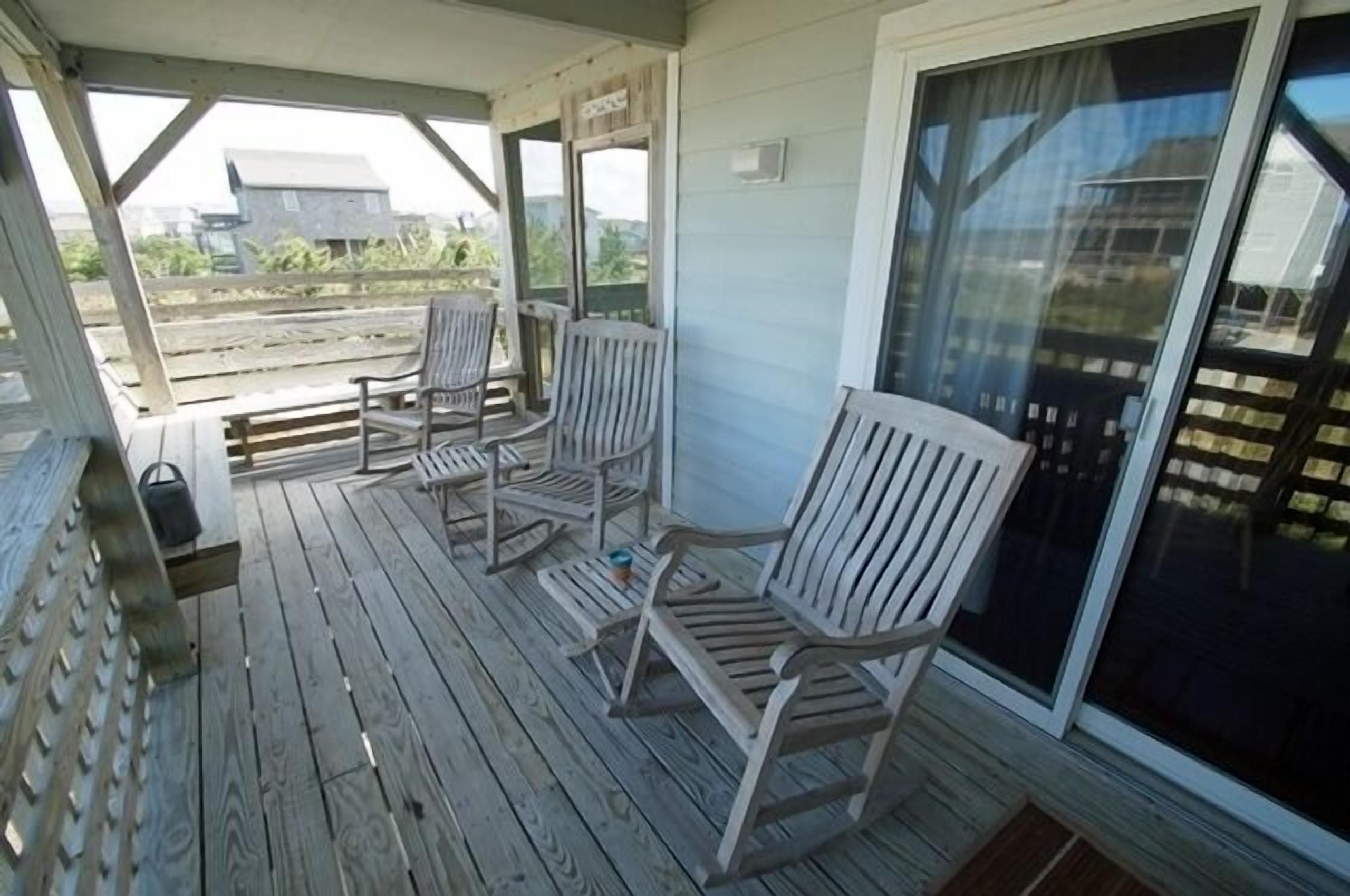 Een overdekt terras met houten terrasvloer, drie houten stoelen en een schuifdeur met glas.