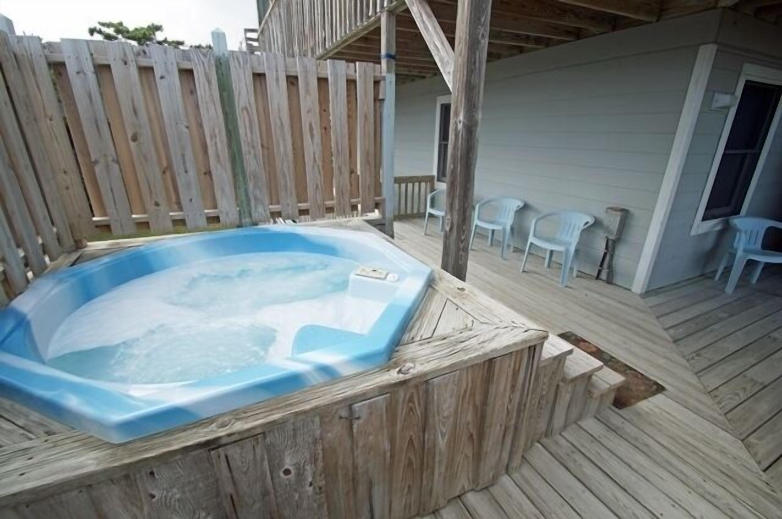 A wooden deck with a hot tub, a covered seating area, and a white building in the background.