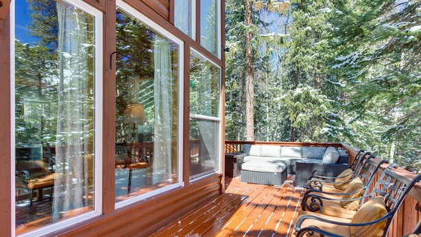 Una terraza de madera con mobiliario exterior y amplios ventanales con vistas a un área boscosa.