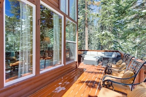 Una terraza de madera con mobiliario exterior, ventanas amplias y vistas a árboles cubiertos de nieve.