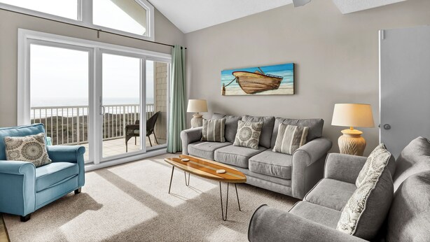 Un vaste salon doté d’une grande porte-fenêtre coulissante donnant sur un balcon, d’un ensemble de canapés gris, d’une table basse en bois et d’un tableau représentant un bateau accroché au mur.