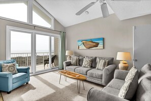 Een ruime woonkamer met een grote schuifpui naar een balkon, een grijze zithoek, een houten salontafel en een schilderij van een boot aan de muur.