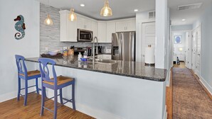 Una cocina moderna con armarios blancos, electrodomésticos de acero inoxidable y encimera de granito. Hay dos taburetes azules y lámparas colgantes sobre la isla.