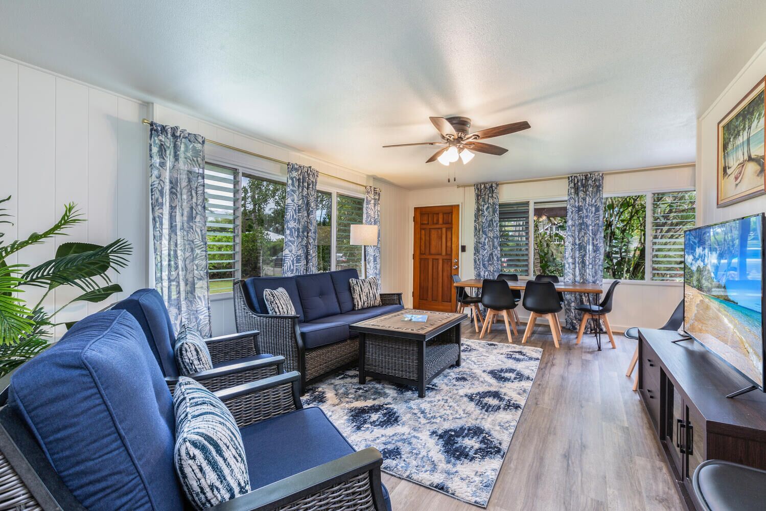 A modern living room with a sofa, chairs, a coffee table, and a TV. There is a ceiling fan, a door, and windows with curtains.