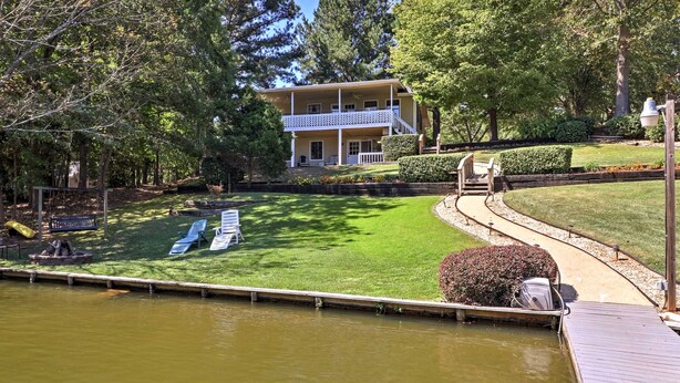 Una propiedad junto al lago con una casa de dos pisos, un muelle y un césped con sillas de descanso.