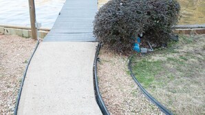 Un sendero de concreto lleva a un muelle de madera sobre aguas tranquilas, con un arbusto y una farola cerca.
