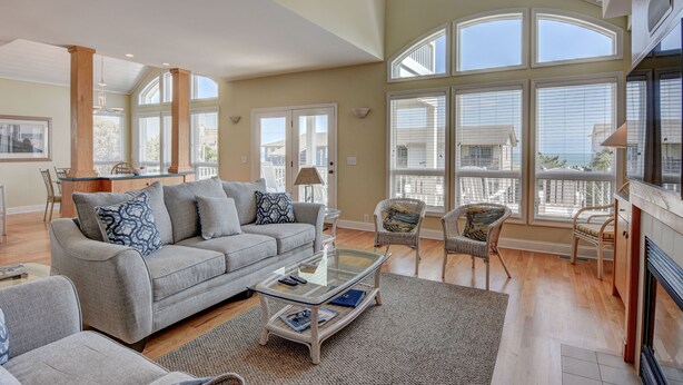 A spacious living room with a grey sofa, a glass coffee table, wicker chairs, and large windows offering a view of the outside. - Schmid – Very spacious & elegant home with outstanding ocean and sound views (Wrightsville Beach)