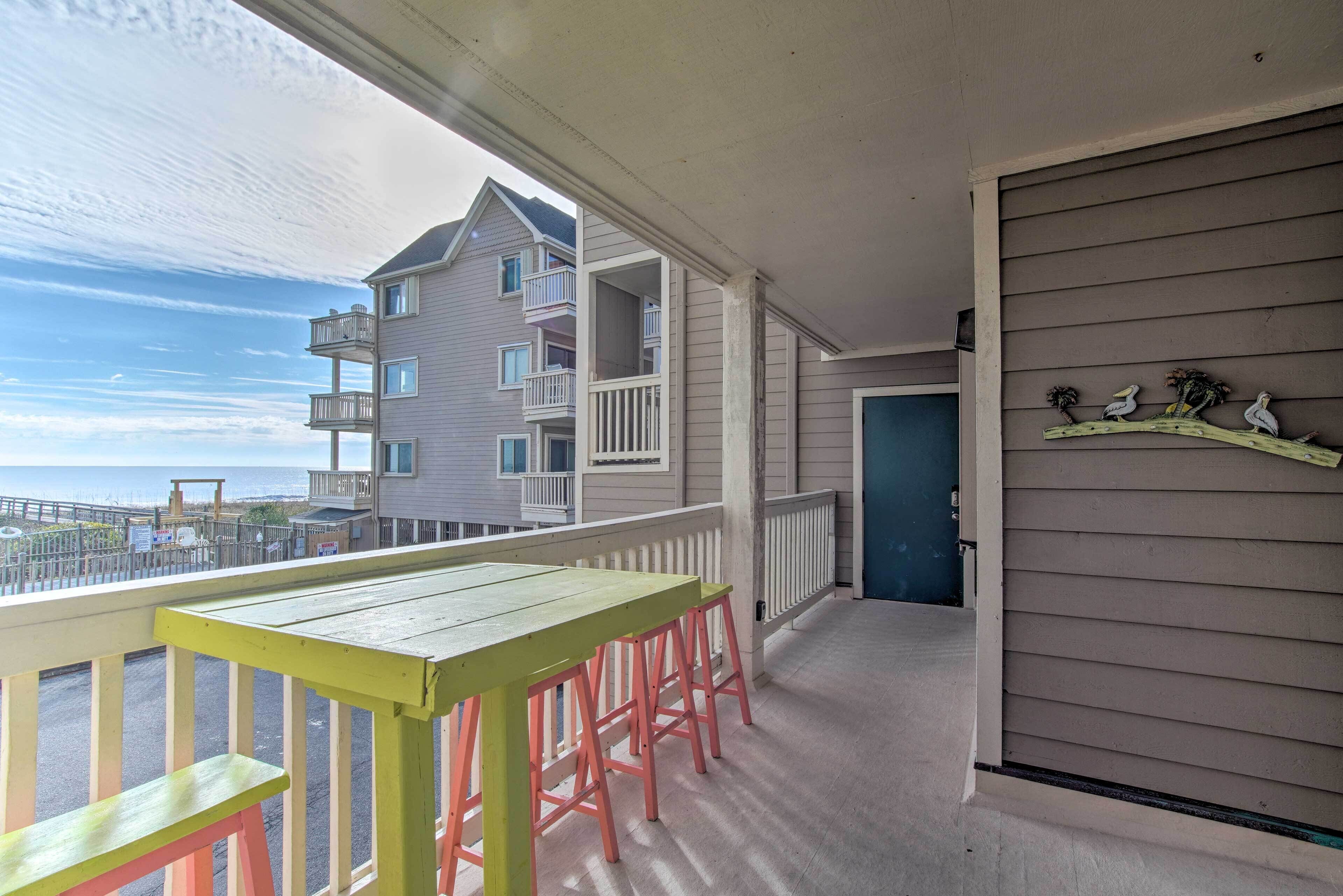 A balcony with a wooden table and pink stools, overlooking a building with multiple balconies and a view of the sea.