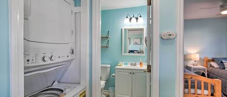 A compact bathroom with a washing machine, a toilet, a sink, and a bed in the adjacent room.