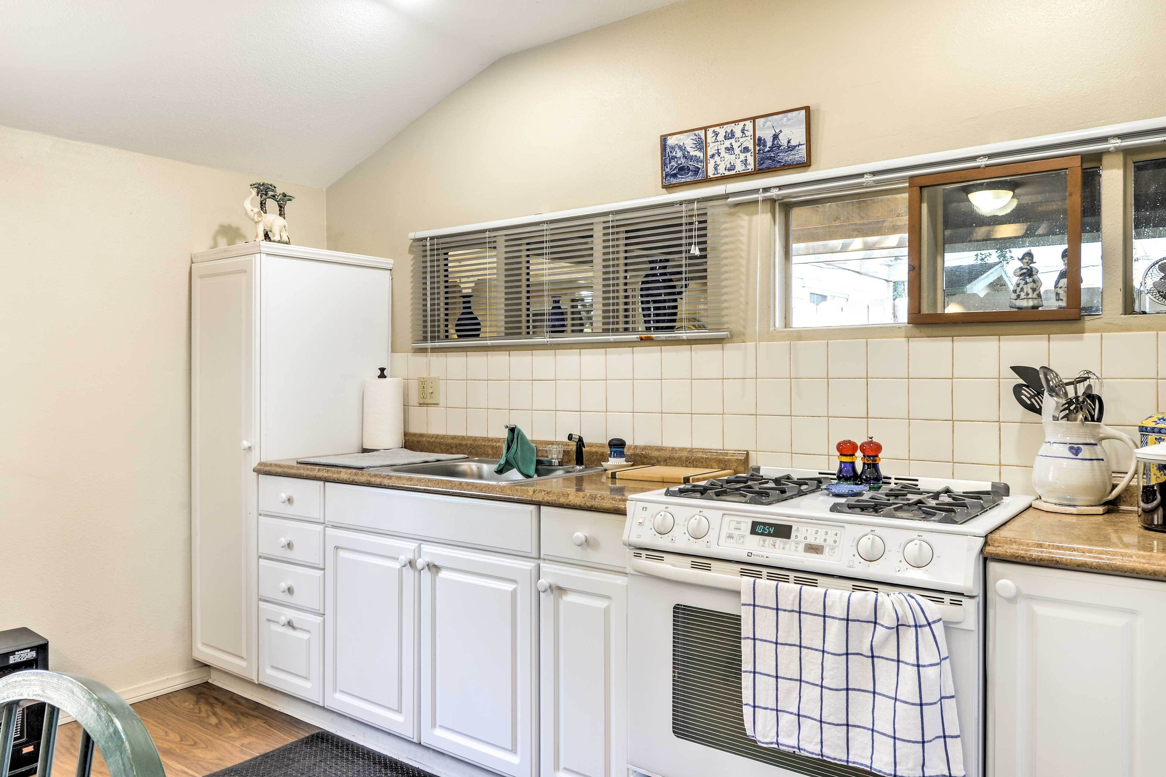 Una cocina con gabinetes blancos, una estufa, un lavamanos y un ventanal con persianas.