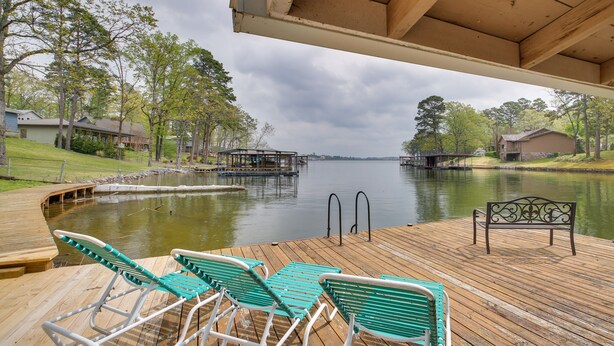 Een houten terras met lounge fauteuils en een bankje met uitzicht op een rustig meer, omringd door bomen en huizen.